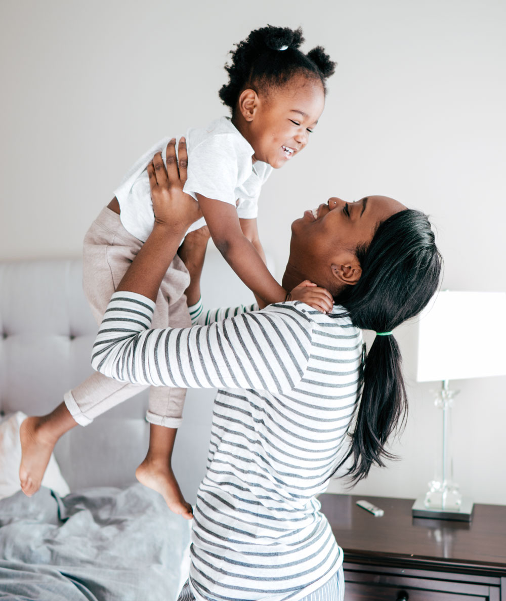 Mother lifting smiling toddler in bedroom.